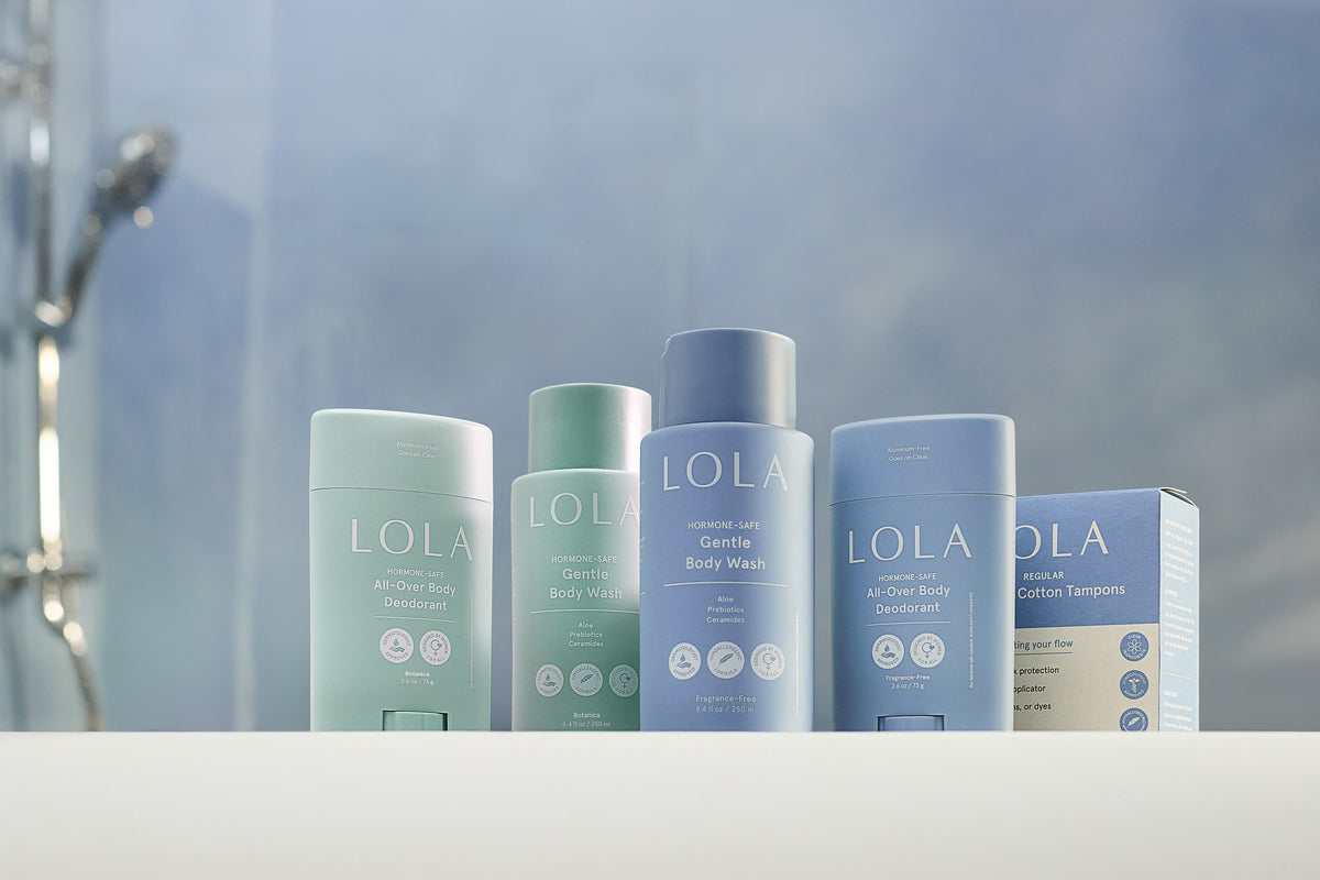 Five LOLA body care products arranged on a white ledge against a softly blurred shower-themed background. The products include body wash, body deodorant in pastel blue, mint green, and light blue packaging.